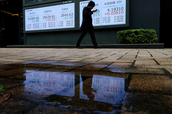 A man is silhouetted, looking down, as he walks past a brightly lit board showing key indicators for stocks and bonds.